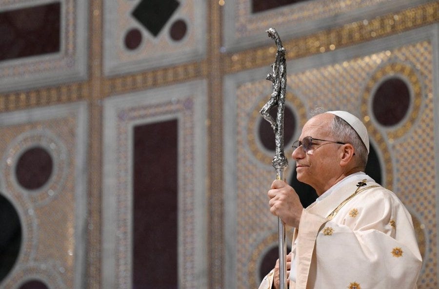 Pope Leo wearing a white and gold embroidered robe, holding a silver staff with a cross design. The background features ornate, colorful mosaic panels with red, gold, and white patterns.