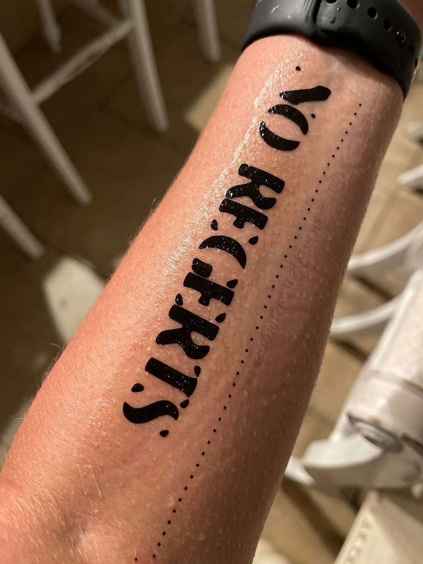 Close-up photo of someone's forearm with a temporary tattoo reading 'NO REGERTS' in black letters, with what appears to be a bike wheel visible in the background.