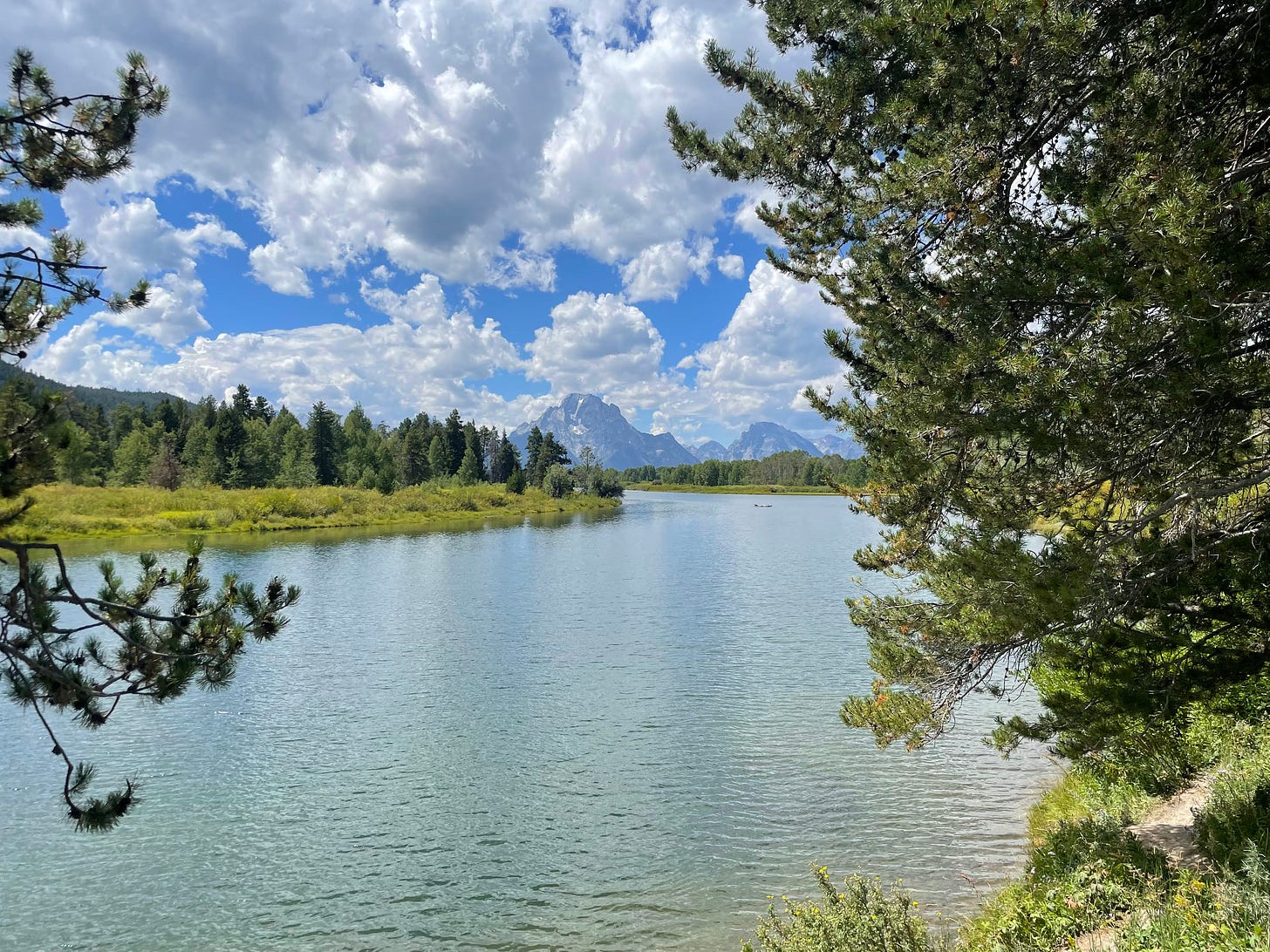 A blue lake is shown, with evergreens on either side and snow-capped mountains in the background. In the blue sky are several puffy white clouds. A blue lake is shown, with evergreens on either side and snow-capped mountains in the background. In the blue sky are several puffy white clouds.