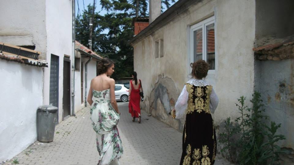 Three women walk away from the camera down a street. They are all in floor length dresses, the woman on the far right in a white dress with a traditional sleeveless maroon and gold dress over the top Three women walk away from the camera down a street. They are all in floor length dresses, the woman on the far right in a white dress with a traditional sleeveless maroon and gold dress over the top
