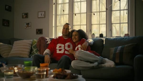 A father and daughter wearing red jerseys and smiling as they watch a football game while sitting on their couch.