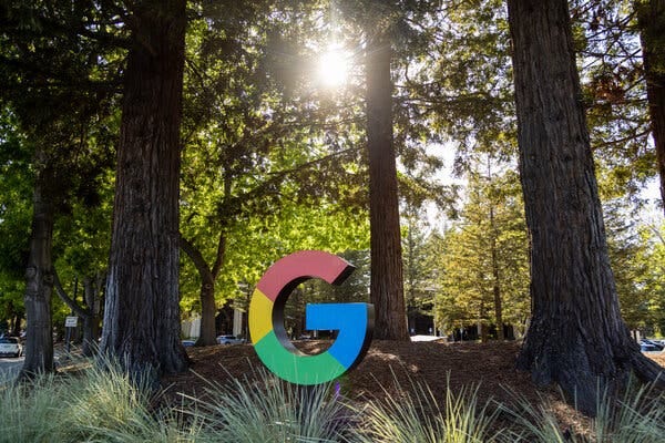 A sculpture of Google’s four-color G logo among tall trees on landscaped grounds.