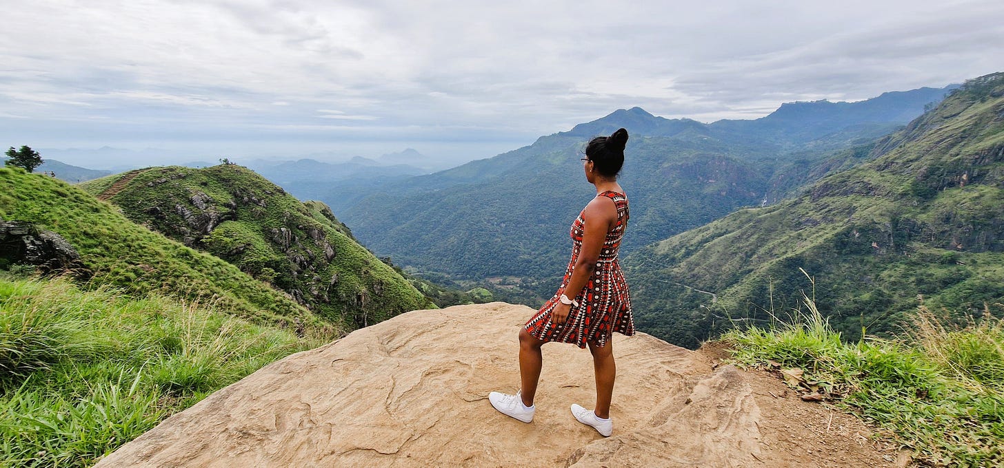 A woman in a red patterned dress standing on a mound, looking out to the mountainous region view below.