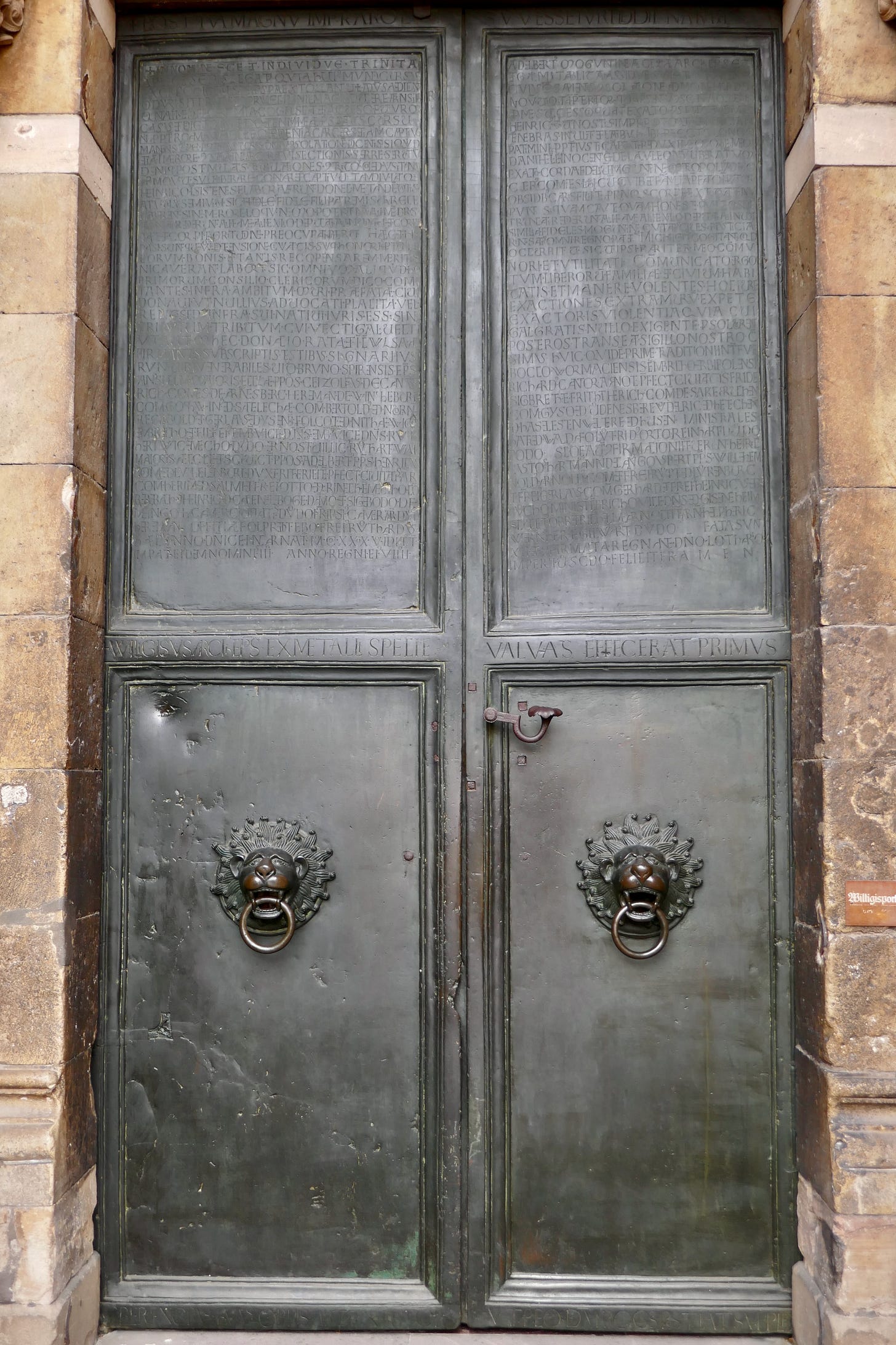 Gray steel doors with four panels and ornate door knockers on each of the bottom two panels, the brick of the structure frames the door