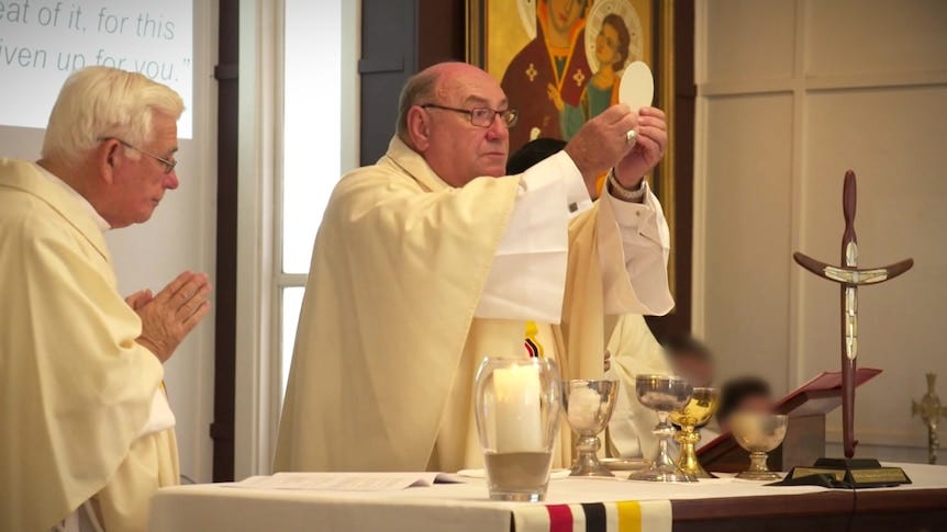 A catholic priest holds up the body of Christ while another priest watches on.