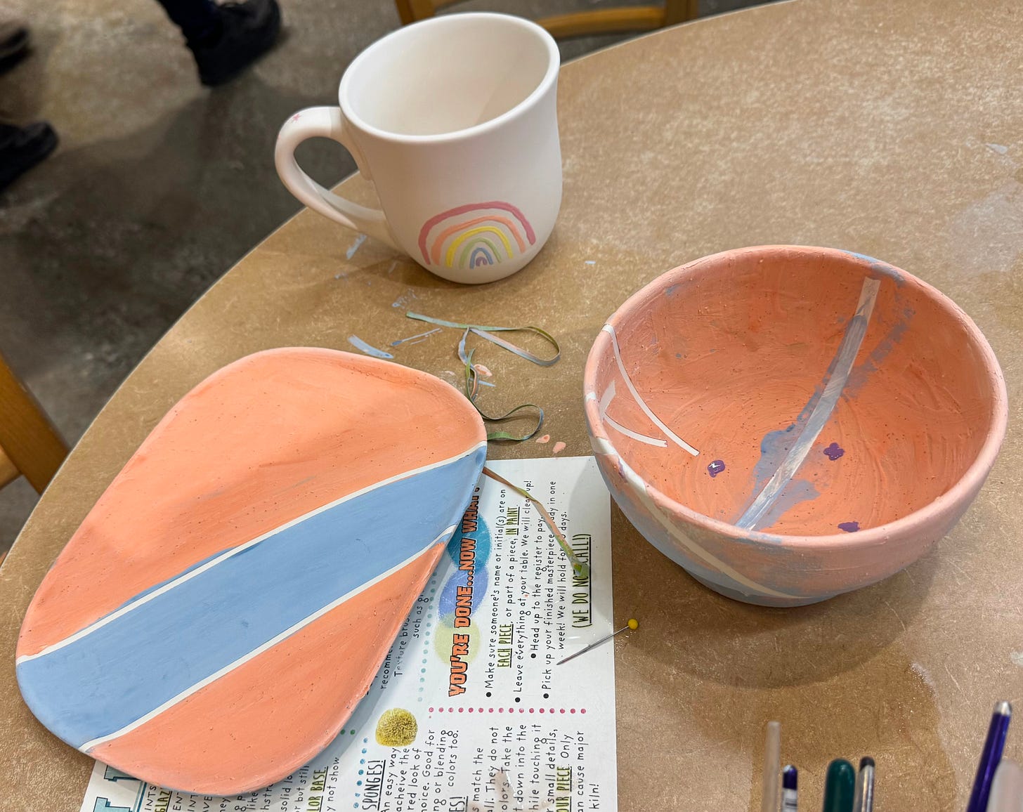 3 pieces of paint your own pottery. one cup with a rainbow painted. one bowl that is orange and blue. one plate with orange and blue stripes.