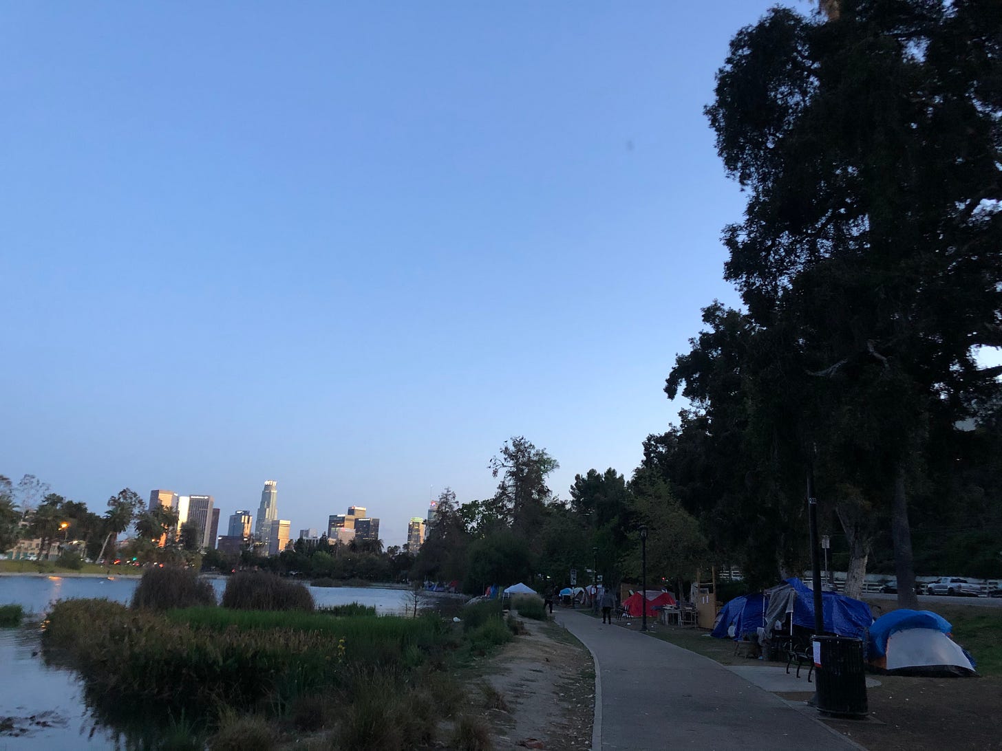 A view of a park lake in the foreground, skyscrapers in the background, and a line of tent shelters on the right.