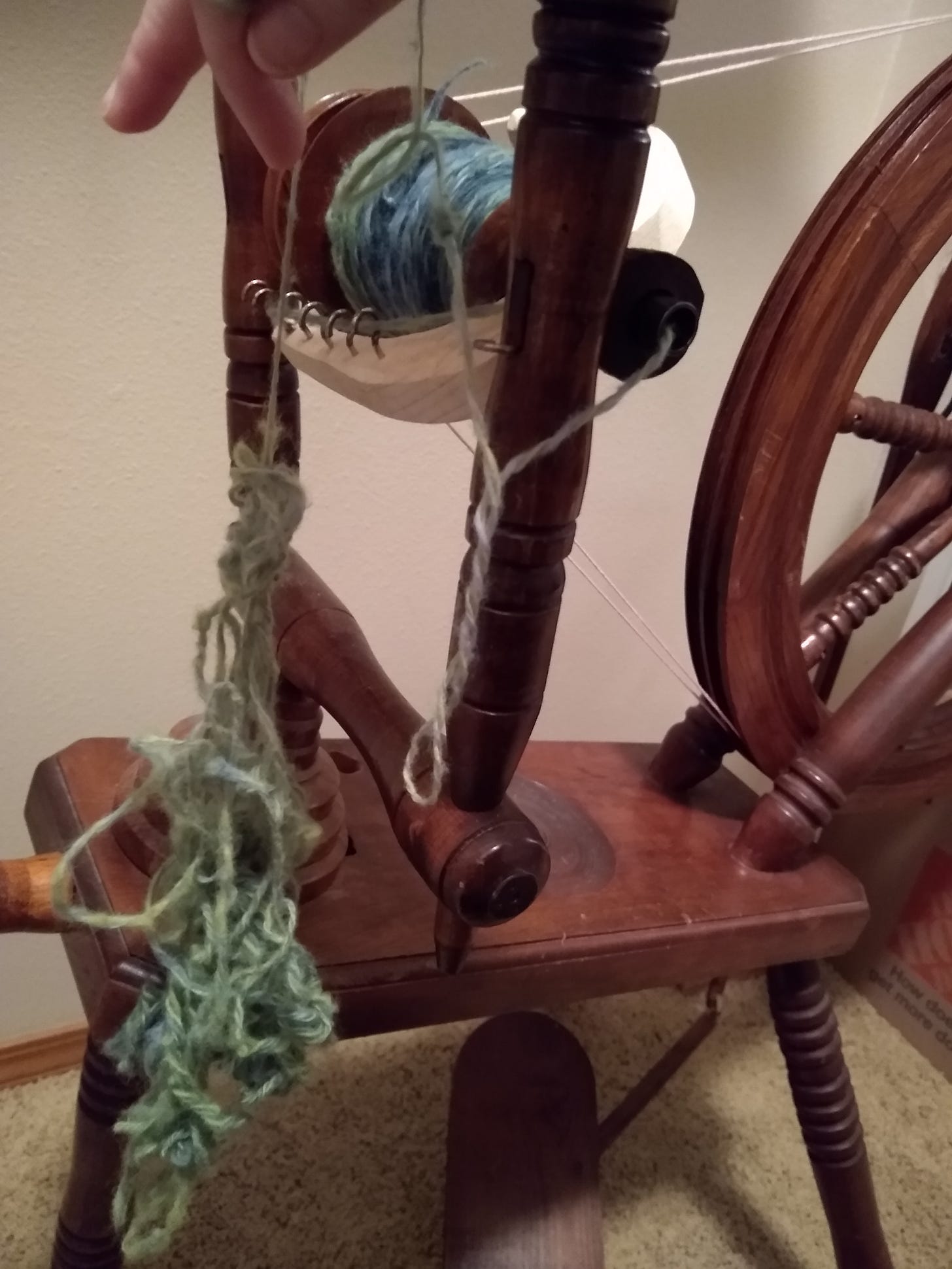 Messy snarl of green yarn hanging in front of spinning wheel, with a mostly nice and neat bobbin of blue and green yarn behind it