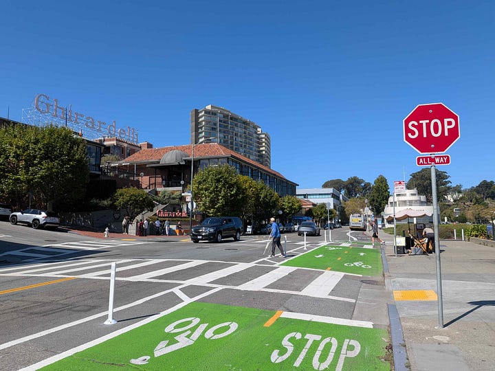 1: Bikeway along the waterfront, the Marin headlands is seen in the distance, 2:Bikeway in front of Ghirardelli Square. Bikeway is painted green. A man is cross the street.