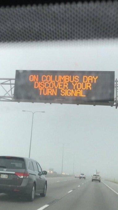 A rainy highway scene shows a large overhead electronic sign displaying On Columbus Day discover your turn signal in yellow text on black background, supported by metal framework with streetlights nearby. A gray SUV is in the foreground viewed from behind through a windshield, with another car trailing on the multi-lane road under overcast skies.