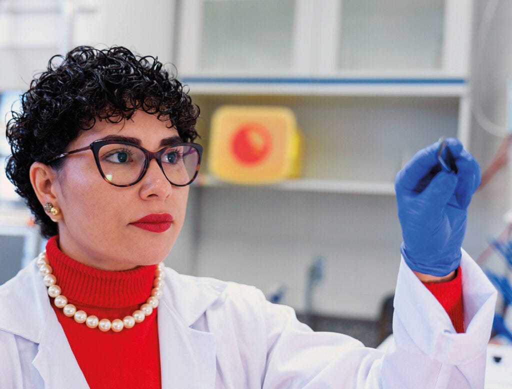 Pessoa usando óculos de armação preta, suéter vermelho, jaleco branco e colar de pérolas, com luva azul na mão direita levantada, em ambiente de laboratório com prateleiras ao fundo contendo frascos e materiais científicos. Pessoa usando óculos de armação preta, suéter vermelho, jaleco branco e colar de pérolas, com luva azul na mão direita levantada, em ambiente de laboratório com prateleiras ao fundo contendo frascos e materiais científicos.