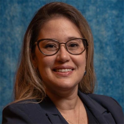 Imagem de uma mulher adulta, de cabelos loiros na altura dos ombros, usando óculos de armação escura. Ela sorri para a câmera e veste um blazer azul-marinho sobre uma blusa preta, com os braços cruzados. Imagem de uma mulher adulta, de cabelos loiros na altura dos ombros, usando óculos de armação escura. Ela sorri para a câmera e veste um blazer azul-marinho sobre uma blusa preta, com os braços cruzados.