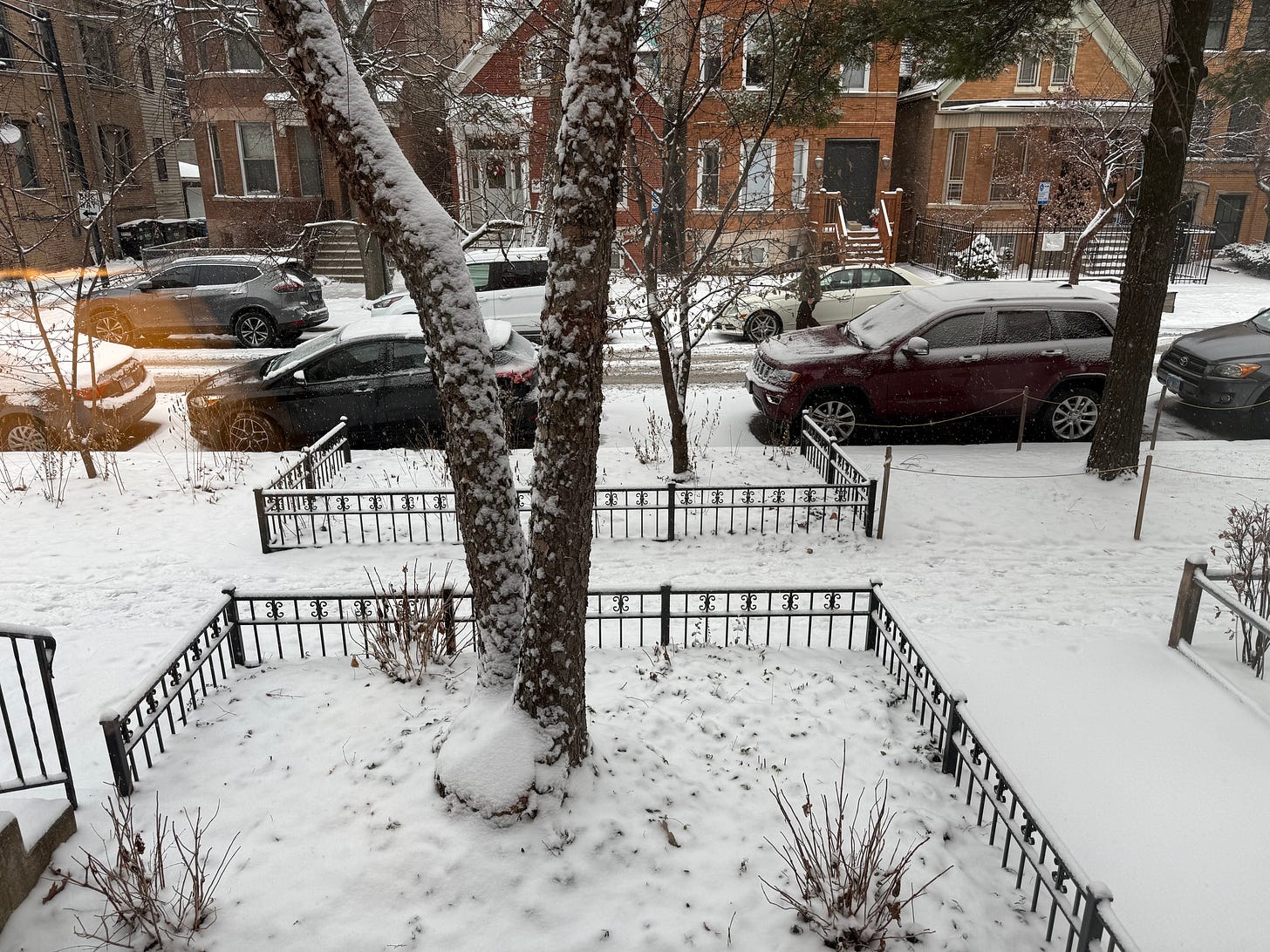 Exterior street scene of trees, car, sidewalks, and roofs covered in a light snow.