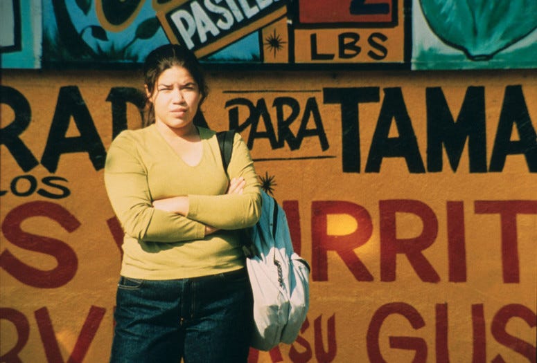 A still from Real Women Have Curves, Patricia Cardoso’s 2000 movie starring America Ferrera. America is standing with her face scrunched, arms crossed, wearing a lime green v-neck pullover and jeans and a backpack slinger over her left shoulder, while waiting in front of a colorful painted wall.