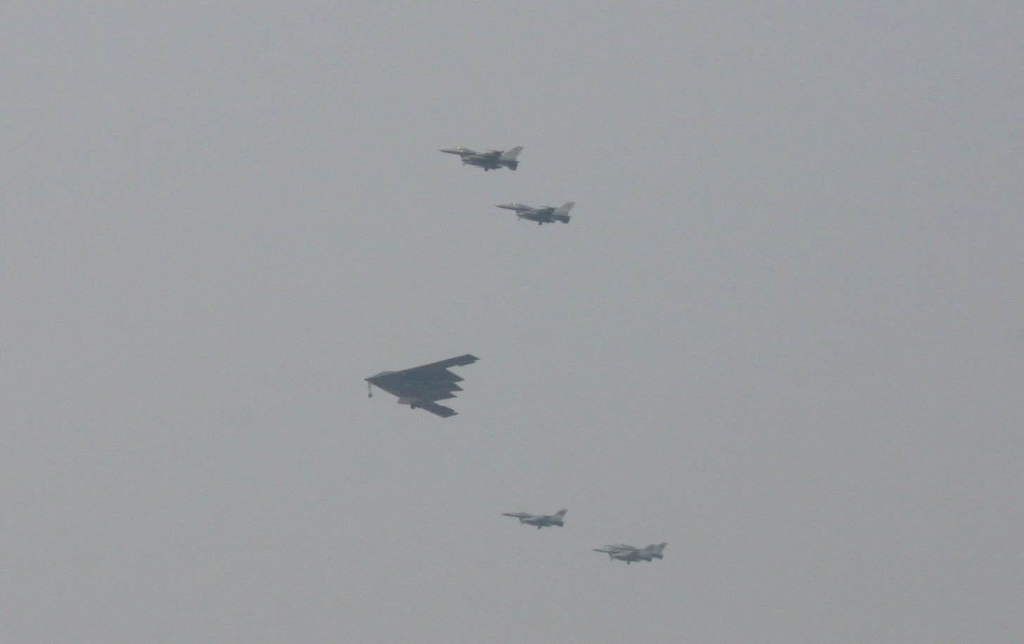A cloudy sky. A B-2 bomber and four F-16s fly in formation over an air base in South Korea. 