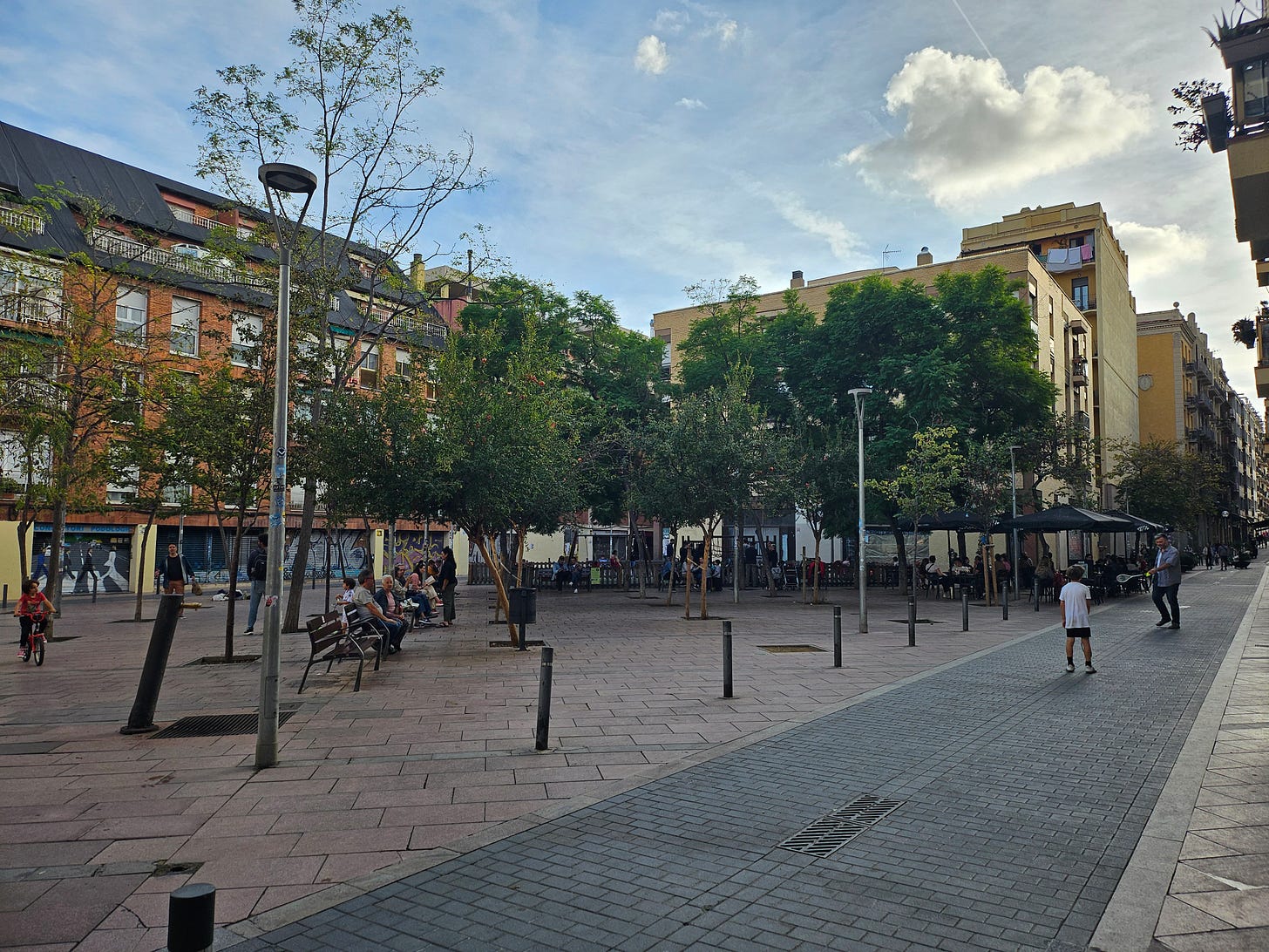 A pedestrianized street in Gràcia, Barcelona, with children playing, people sitting at cafe tables, and planters with greenery.