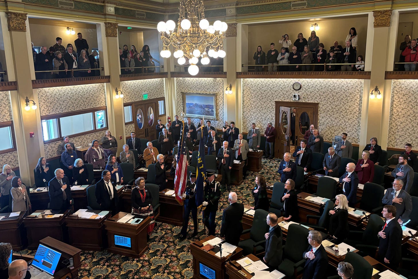 A photo of Montana state senators standing as a two flagbearers carry a U.S. and Montana flag into the room. A photo of Montana state senators standing as a two flagbearers carry a U.S. and Montana flag into the room.