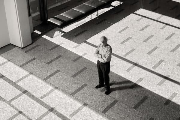A black-and-white image shows a man, wearing trousers and a button-down shirt, standing with his arms crossed in the lobby of a concert hall.