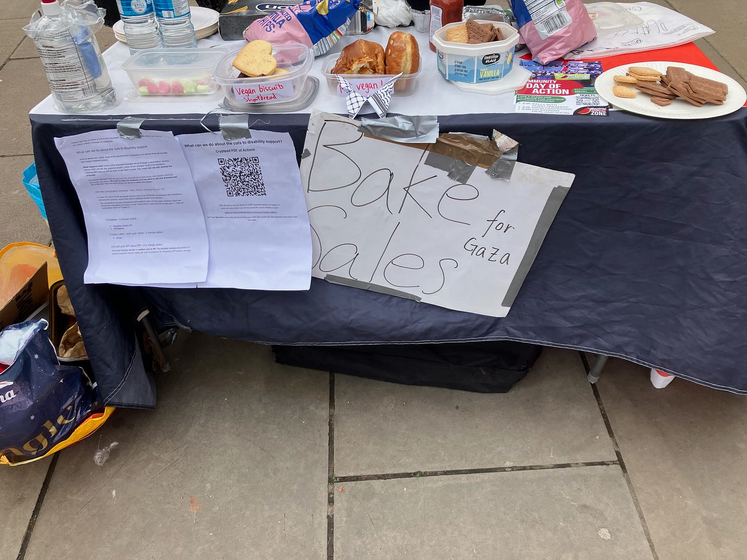 A stall outside on the street that has two signs one saying 'what we can do about the cuts to disability' with a list of actions and one that says 'bake sale for Gaza'