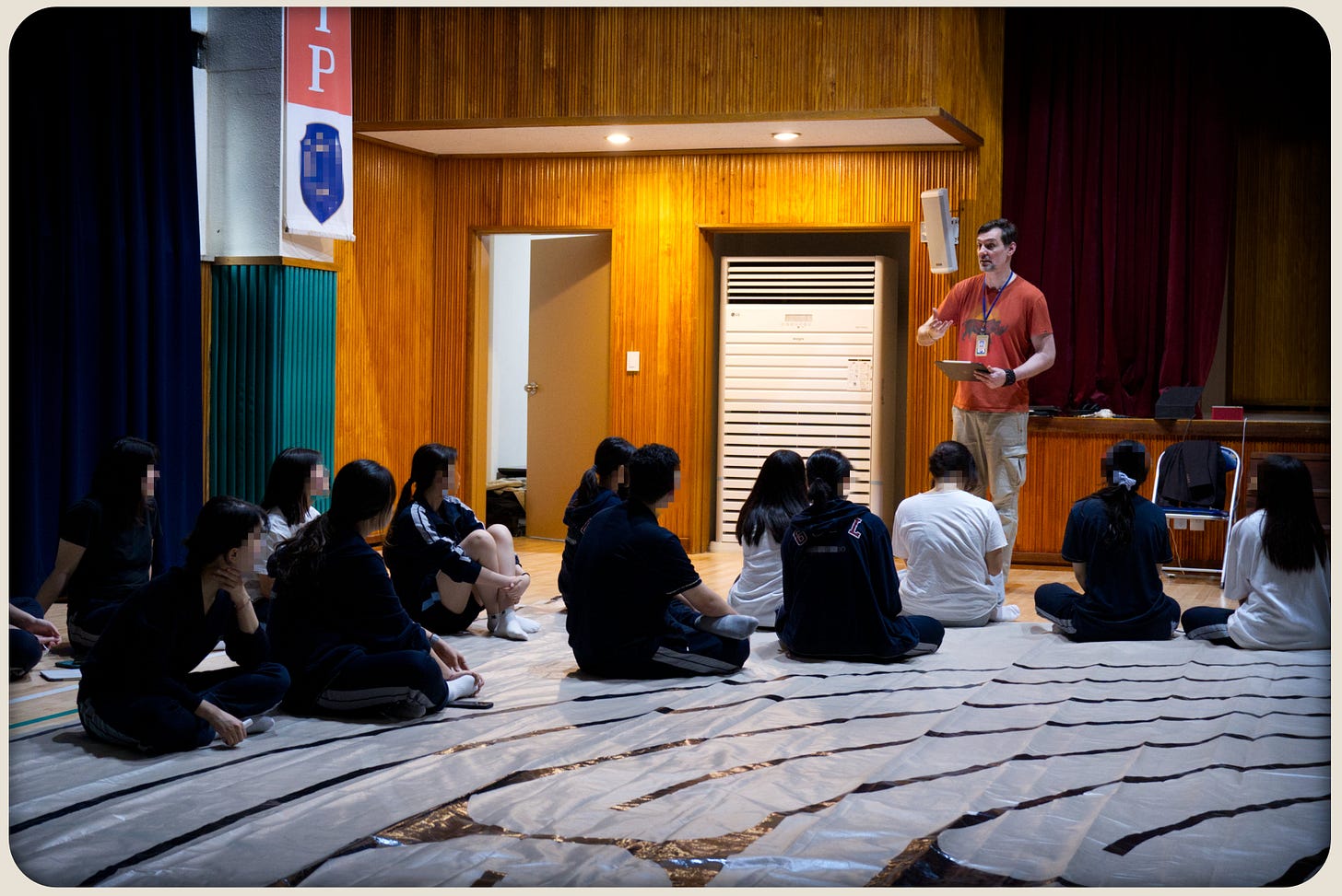 Students seated on a large tarp labyrinth in a school auditorium while a facilitator stands and gives instructions.