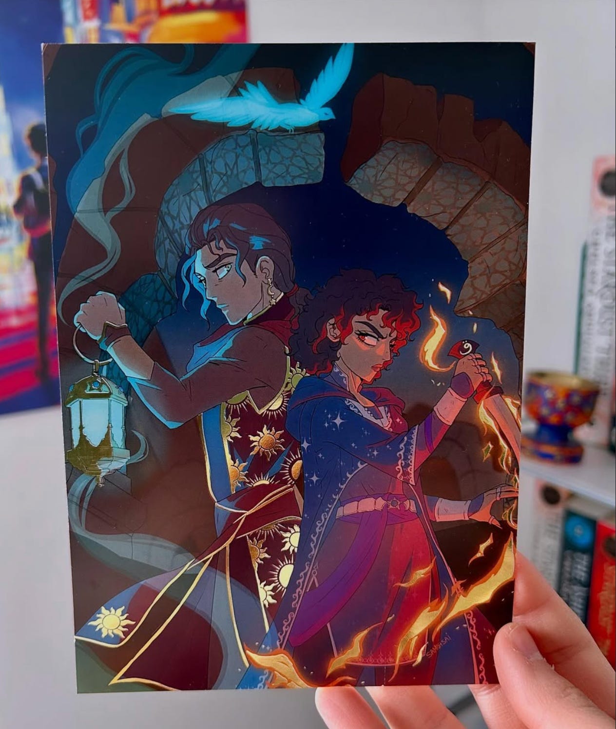 A foiled print featuring Mazen & Loulie from THE ASHFIRE KING. The characters stand back to back against ruins. Mazen holds a lantern with a flaming bird floating above and Loulie holds a flaming blade. The fire around Loulie’s blade, as well as the sunny patterns on Mazen’s vest, are foiled. 