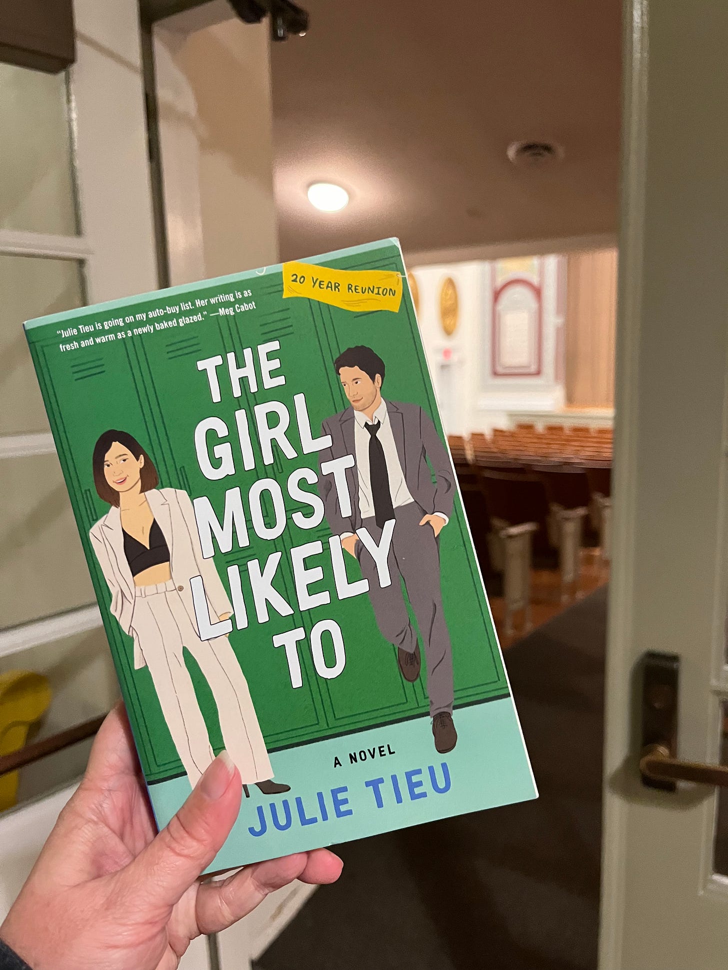 Photo of my hand holding up THE GIRL MOST LIKELY TO by Julie Tieu in front of an auditorium