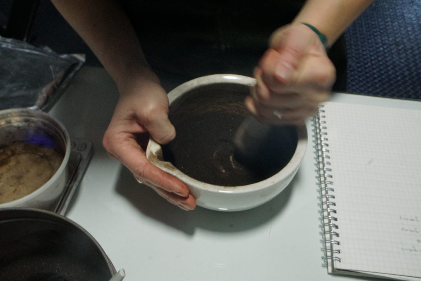 A dark brown powder (asphaltum) is being ground with a pestle in a large white mortar. A notebook rests to the right of the mortar.