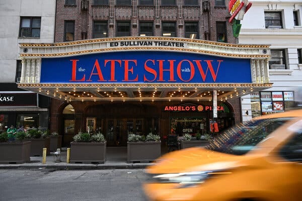 A theater marquee with “Late Show” in big red letters against a blue background. The top of the marquee says, “Ed Sullivan Theater.” A blurred car passes in the foreground.