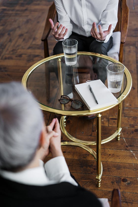 A grey haired man with his legs crossed sitting and listening while another person gestures across from them. On the table is water, glasses, and a pen and paper. A grey haired man with his legs crossed sitting and listening while another person gestures across from them. On the table is water, glasses, and a pen and paper.