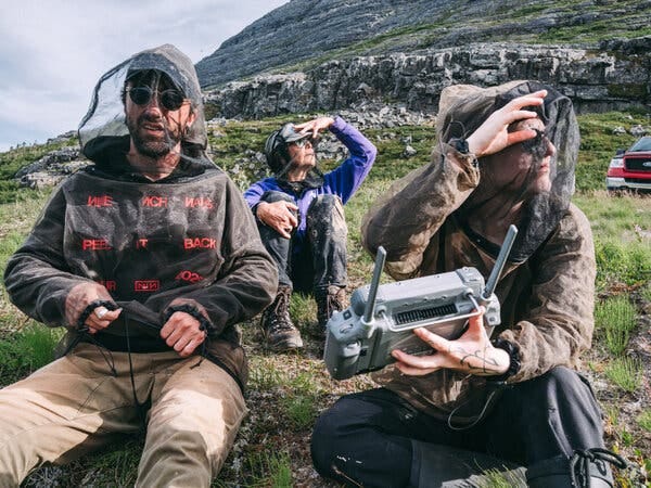 Three people wearing bug nets are seated on rocky terrain. One shields their eyes, another holds a drone remote.