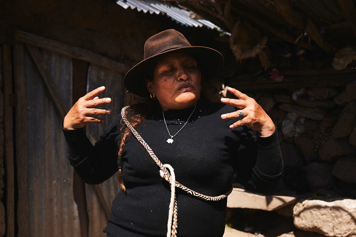 A woman wearing a brown hat and black sweater stands outside with her eyes closed and hands raised near her face, gesturing expressively. Sunlight highlights her face, and rustic wooden and stone structures are in the background.