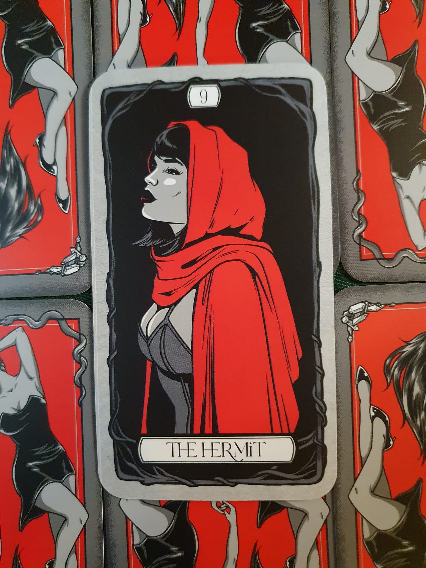 The Hermit tarot card, numbered nine, showing a dark-haired woman wearing a flowing red hooded cloak over a black dress. She looks thoughtfully over her shoulder against a black background, framed by twisting dark branches. The Hermit tarot card, numbered nine, showing a dark-haired woman wearing a flowing red hooded cloak over a black dress. She looks thoughtfully over her shoulder against a black background, framed by twisting dark branches.