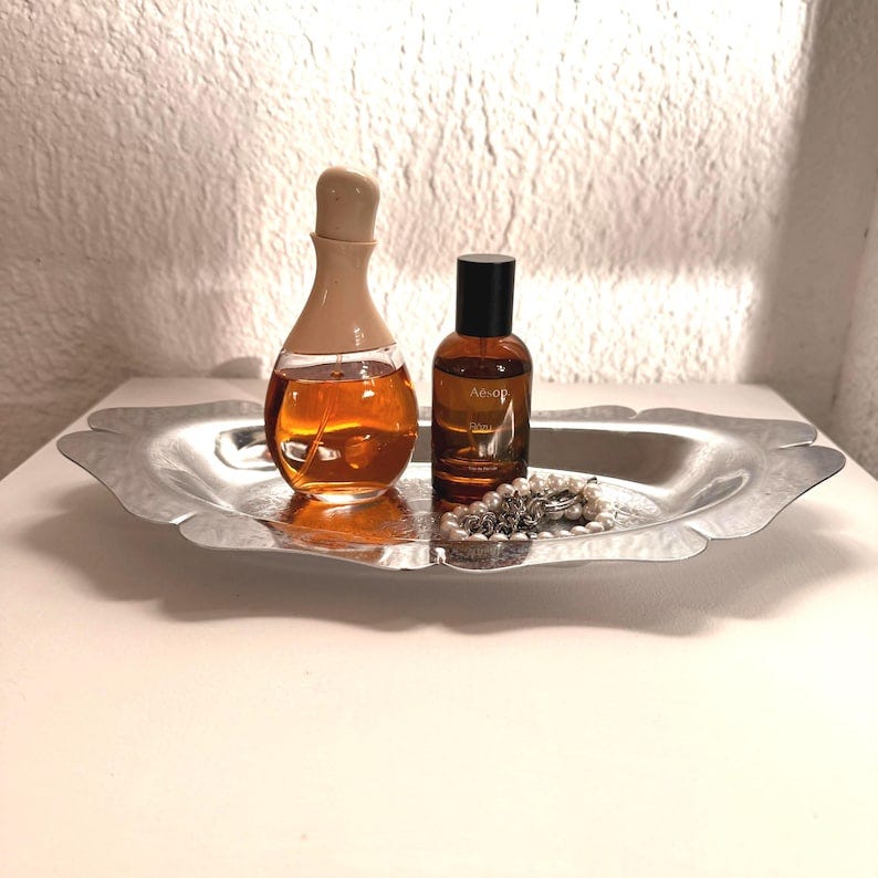 May include: Two bottles of perfume on a silver tray with a pearl necklace. The bottle on the left is clear with a beige cap and the bottle on the right is dark amber with a black cap. The bottle on the right has the text "Aesop" on the label.