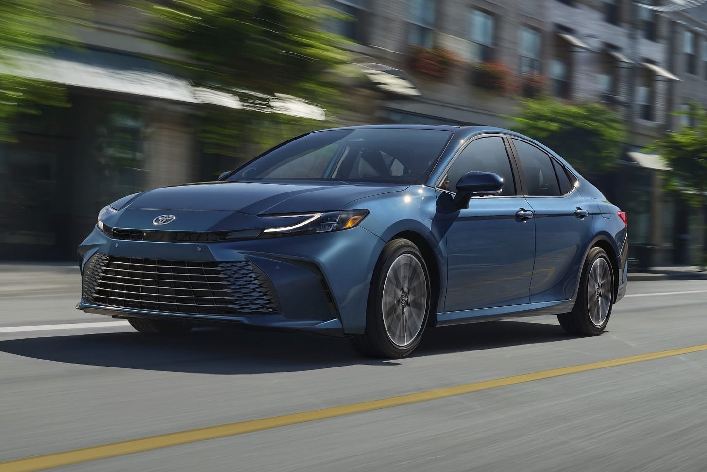 A blue 2025 Toyota Camry XLE hybrid drives on a tree-lined city street on a sunny day.