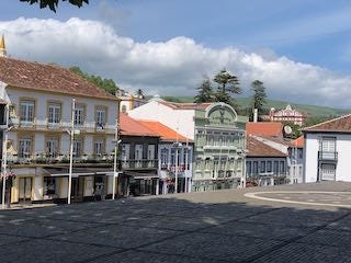 Photos of black rocks in a frothy ocean, a stone wall in front of fields and a village, a town plazaa with colorful buildings and orange tile roofs.