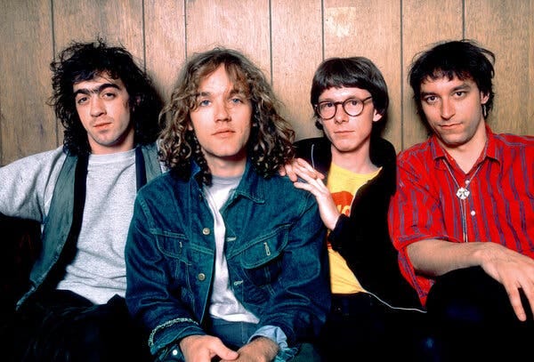 A color photograph of the band R.E.M., four young men seated against a wood-paneled wall. Their haircuts and clothes indicate that the photograph was taken in the 1980s.