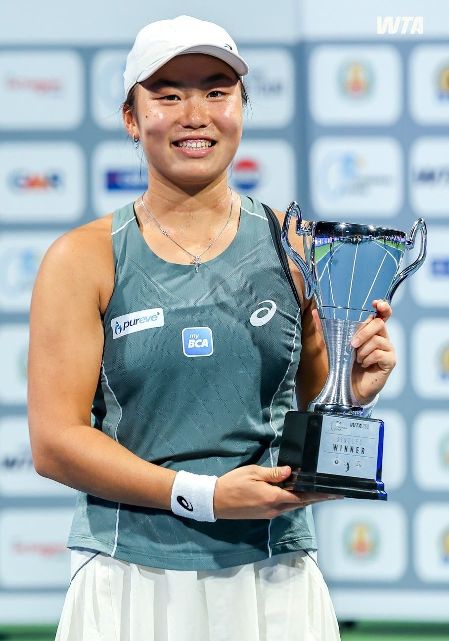 Indonesian tennis player Janice Tjen smiles while holding her winner’s trophy after clinching the 2025 WTA Chennai Open singles title. She wears a gray Asics outfit and white cap, standing against a sponsor backdrop featuring the WTA logo.