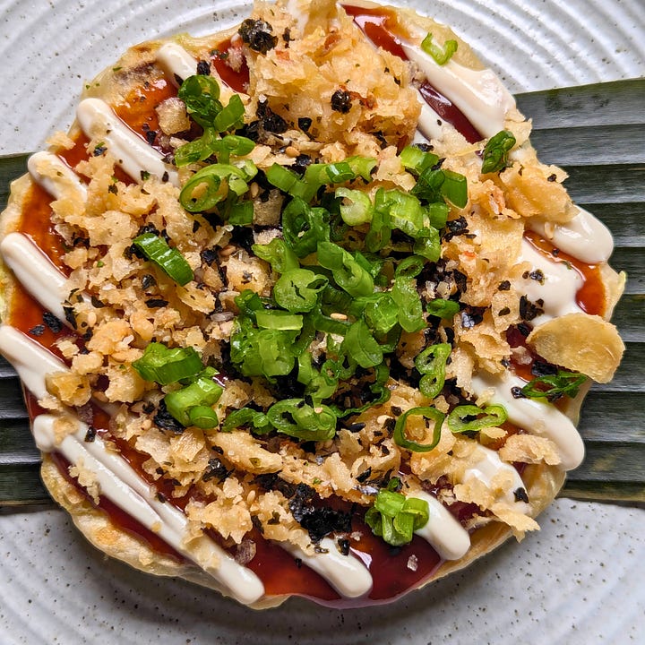 Two finished okonomiyaki pancakes, each topped with okonomiyaki sauce, vegan mayo, and different garnishes.