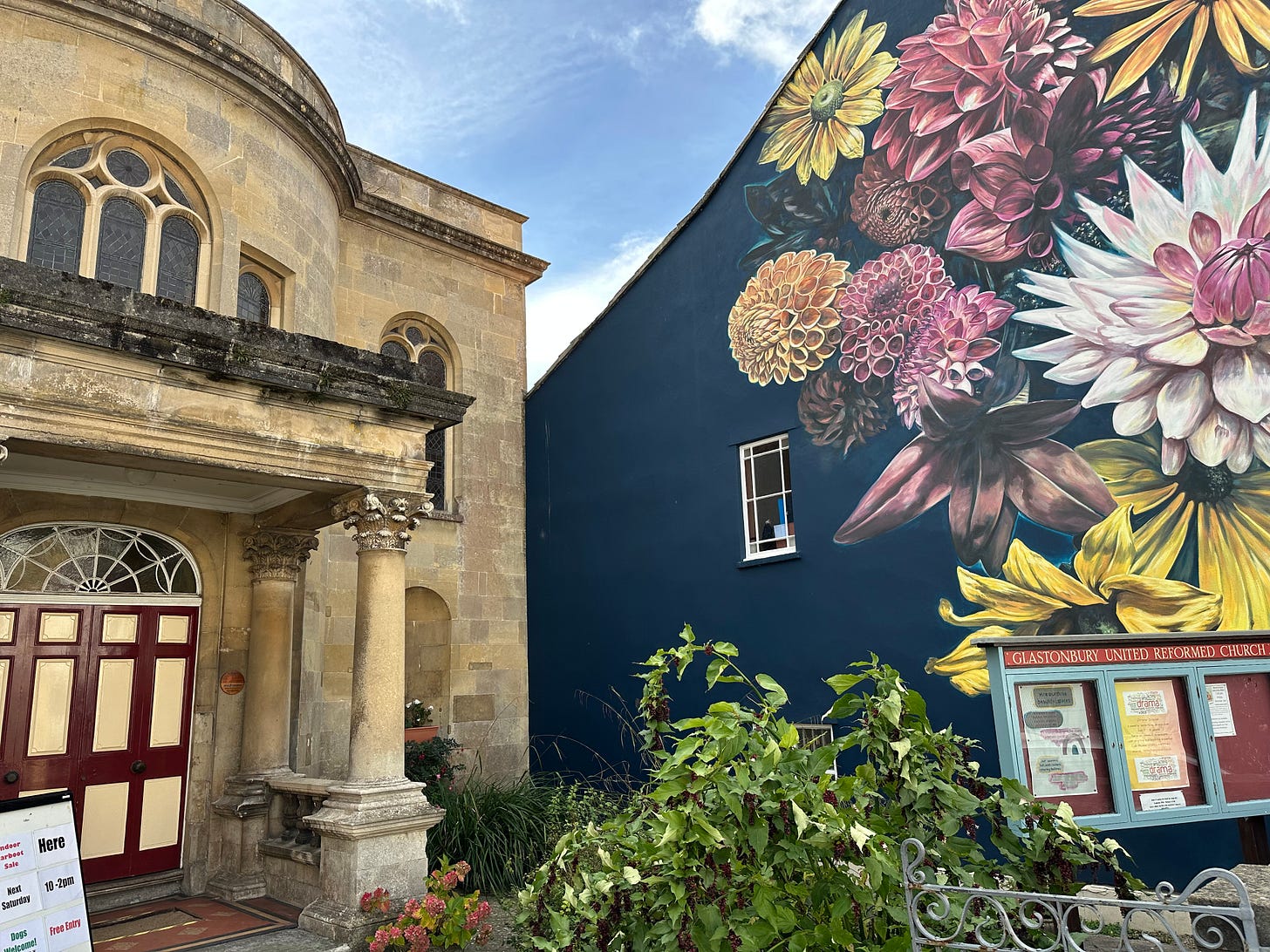 The front of Glastonbury United Reformed Church with its stone columns and arched windows, beside a neighbouring building decorated with a vibrant mural of large flowers.
