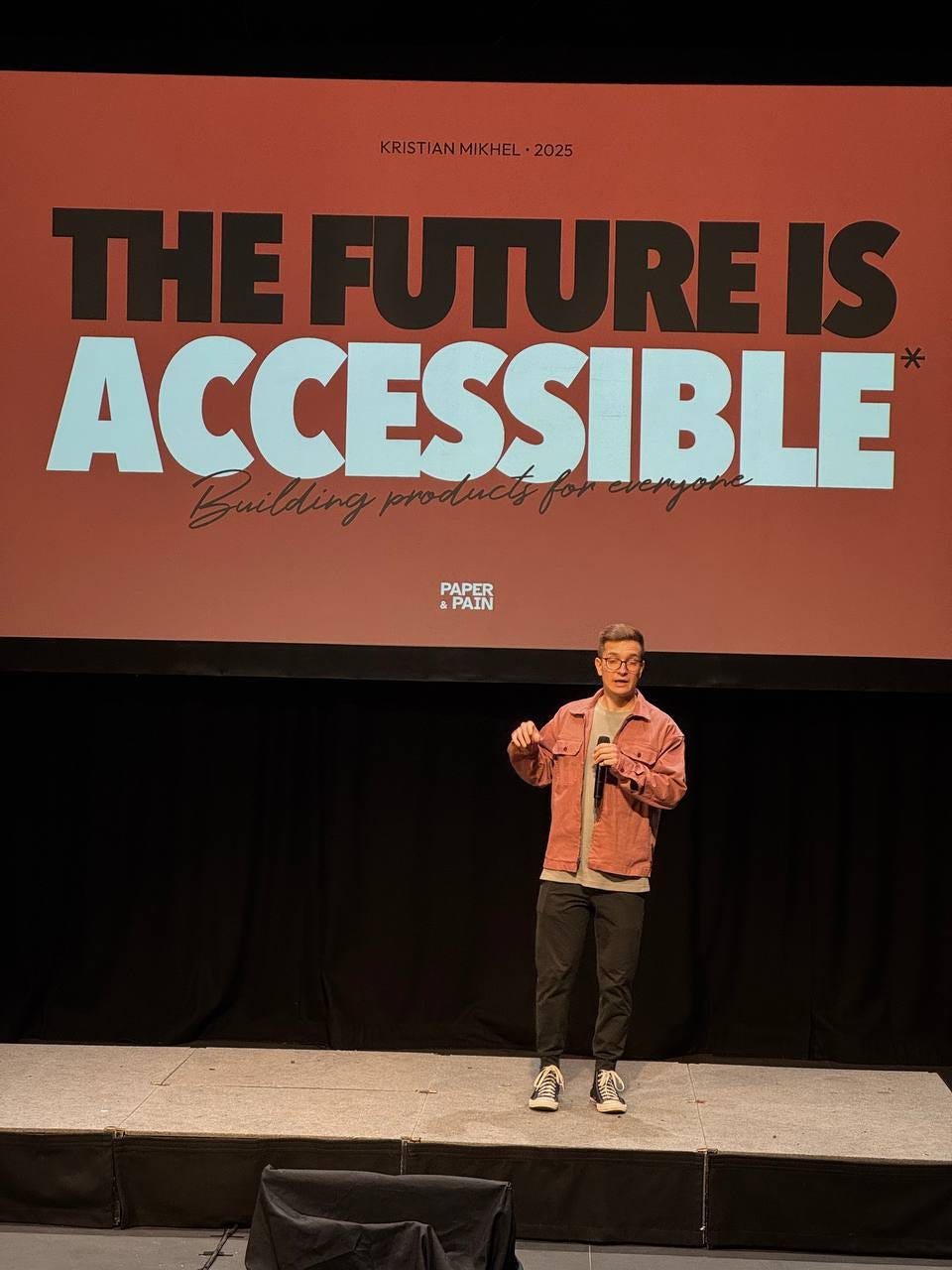 A young handsome man on stage in front of the massive screen backdrop. Caption at the top of the slide: "Kristian Mikhel, 2025". Title: "The future is accessible. Building products for everyone". By Paper and Pain.