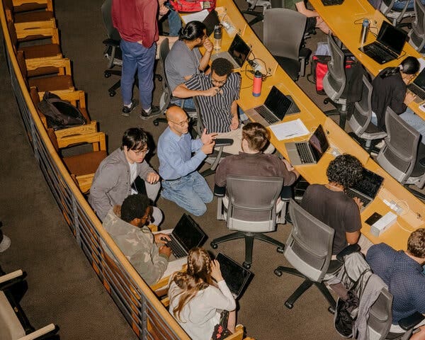 Jeremy Avigad, in jeans and a blue Oxford shirt, kneels amid a dozen students who sit in a lecture hall in front of laptops.