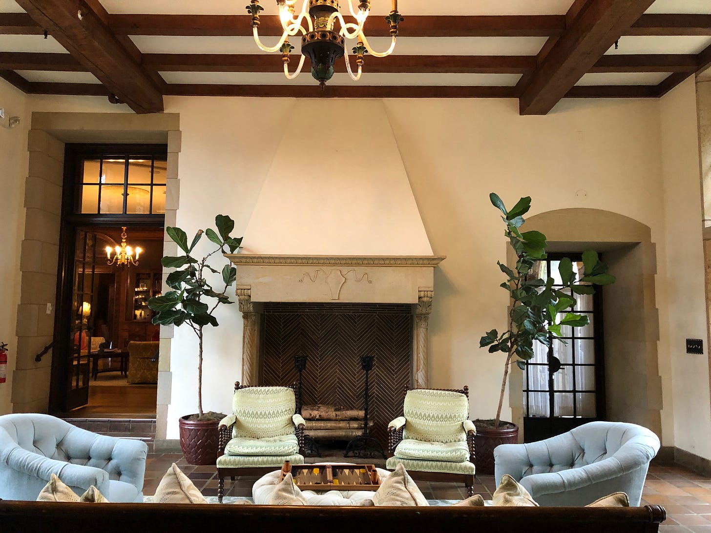A wooden beamed ceilinged room with four chairs around a low table. A fireplace with potted plants around it. Two doorways at the back.