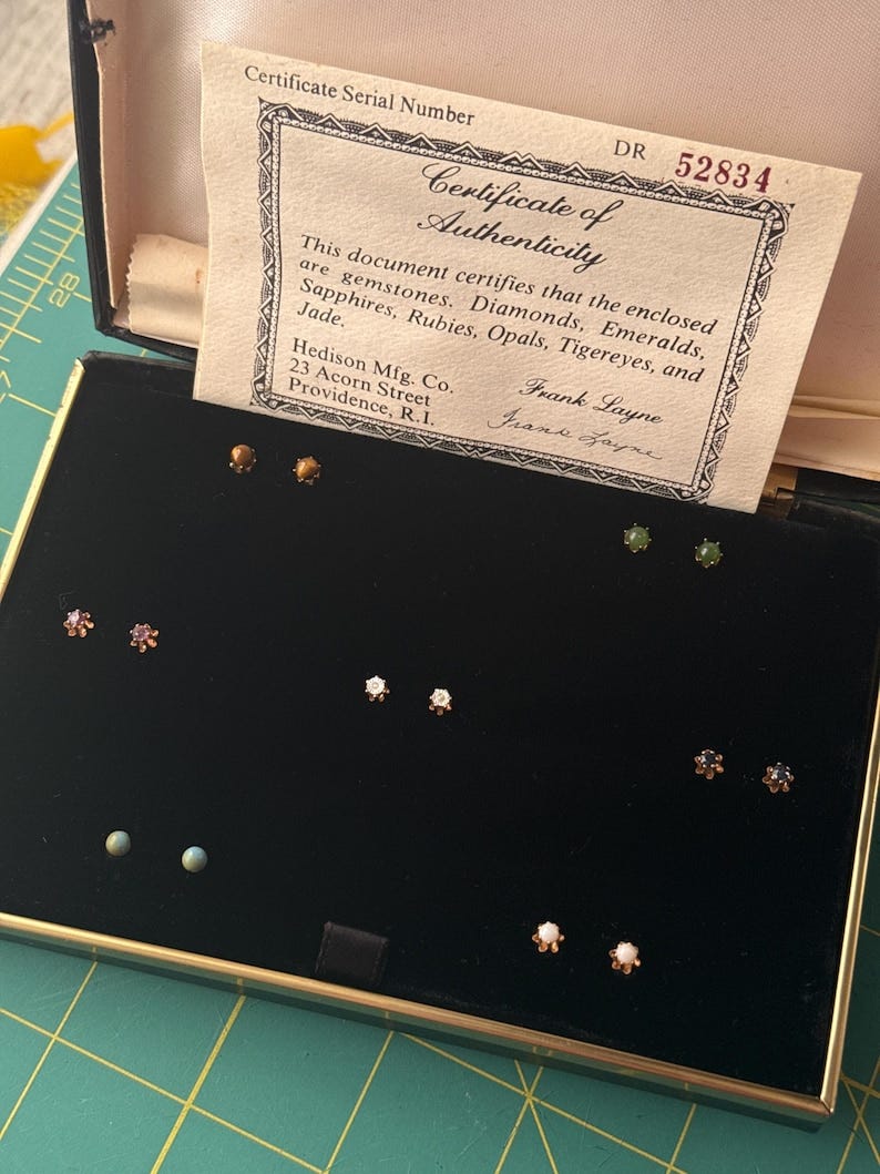 May include: An open jewelry box with a black velvet interior, displaying various gemstone stud earrings. A certificate of authenticity is visible, listing gemstones like diamonds, emeralds, and sapphires. The box has a gold-colored frame.