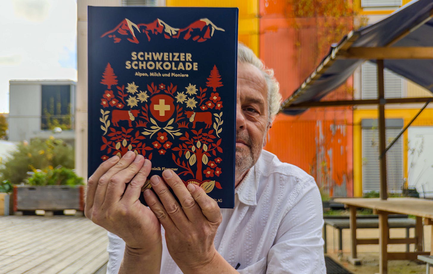 Der Schweizer Ernährungshistoriker Dominik Flammer sitzt im Freien und hält sein neues Buch «Schweizer Schokolade – Alpen, Milch und Pioniere» vor sich, sodass das kunstvoll illustrierte Cover mit Alpenmotiven, Kuh und Schweizer Kreuz im Mittelpunkt steht. Flammer blickt halb hinter dem Buch hervor, im Hintergrund sind farbige Gebäudefassaden zu sehen. Der Schweizer Ernährungshistoriker Dominik Flammer sitzt im Freien und hält sein neues Buch «Schweizer Schokolade – Alpen, Milch und Pioniere» vor sich, sodass das kunstvoll illustrierte Cover mit Alpenmotiven, Kuh und Schweizer Kreuz im Mittelpunkt steht. Flammer blickt halb hinter dem Buch hervor, im Hintergrund sind farbige Gebäudefassaden zu sehen.
