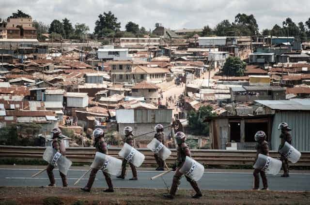People in helmets, carrying truncheons and shields, walk on a road with a dense informal settlement in the background People in helmets, carrying truncheons and shields, walk on a road with a dense informal settlement in the background