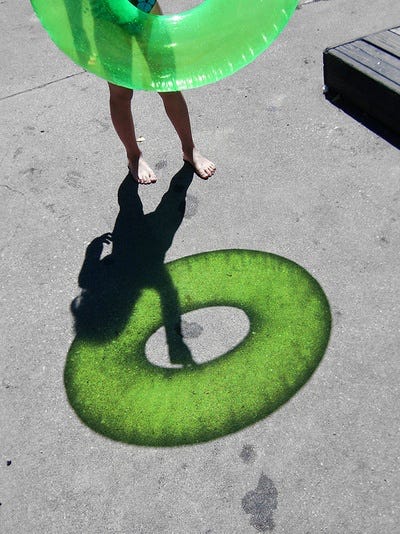 A series of symbols and signs including a the shadow of a green rafting tube, a lady bug on a four leaf clover, a street full of green stoplights, green birds on a line, a white arrow painted on the ground with green grass growing through it, a digital clock with the numbers 11:11