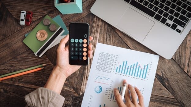 A person using a calculator on their phone with spreadsheets on their desk