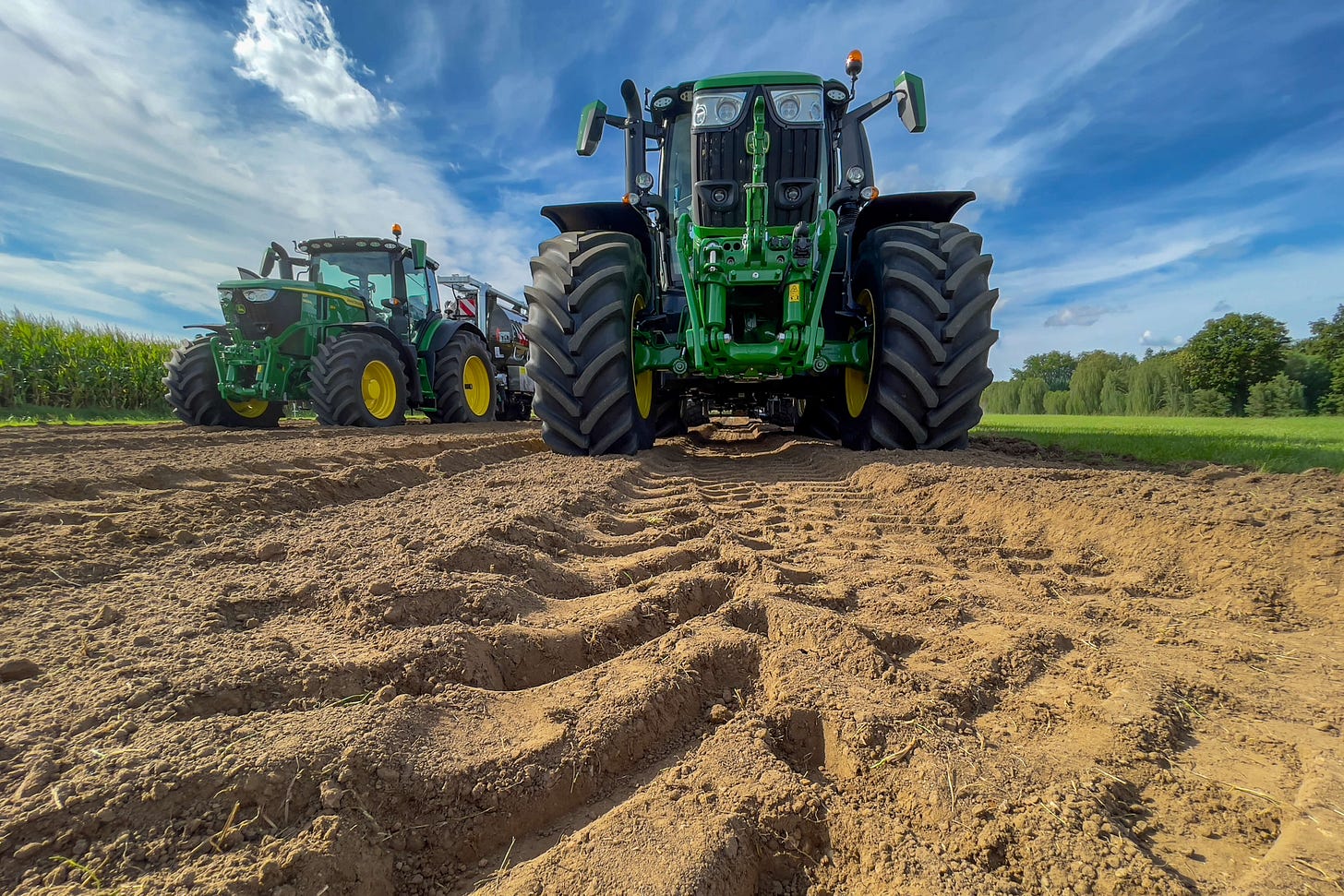 Zwei Gross-Traktoren vom Typ John Deere 6R 250 mit der markentypischen grasgrünen Lackierung und gelben Felgen stehen frontal auf einem Acker; im Vordergrund sind tiefe Reifenspuren und aufgeworfener Boden sichtbar, im Hintergrund Felder, Bäume und ein blauer Himmel mit Wolken.