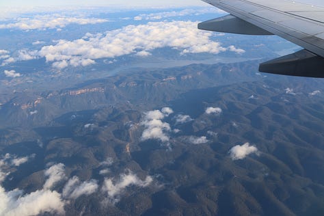 Top left - from the airplane window through wisps of cloud to rolling hills. Top middle, from the airplane window, big highway lanes disappear into the distance as the plane banks to land. Top right - bicycle leaning against a pole at a roadside stall. Middle left, small grey house with hedge. Middle middle, dirt road town with two storey shops, wires overhead. Middle right. City square with man leaning looking at the camera with big old building in the background. Cobblestone streets. Bottom left. Close up of bicycle at roadside stop. Road visible with a big blue lorry speeding past. Bottom middle. Most of an orange building with a curved roof and supporting tower wall, graffiti and run down with weeds in the foreground. Bottom right. Big storm clouds over twin railings and city in the background, weeds in the foreground