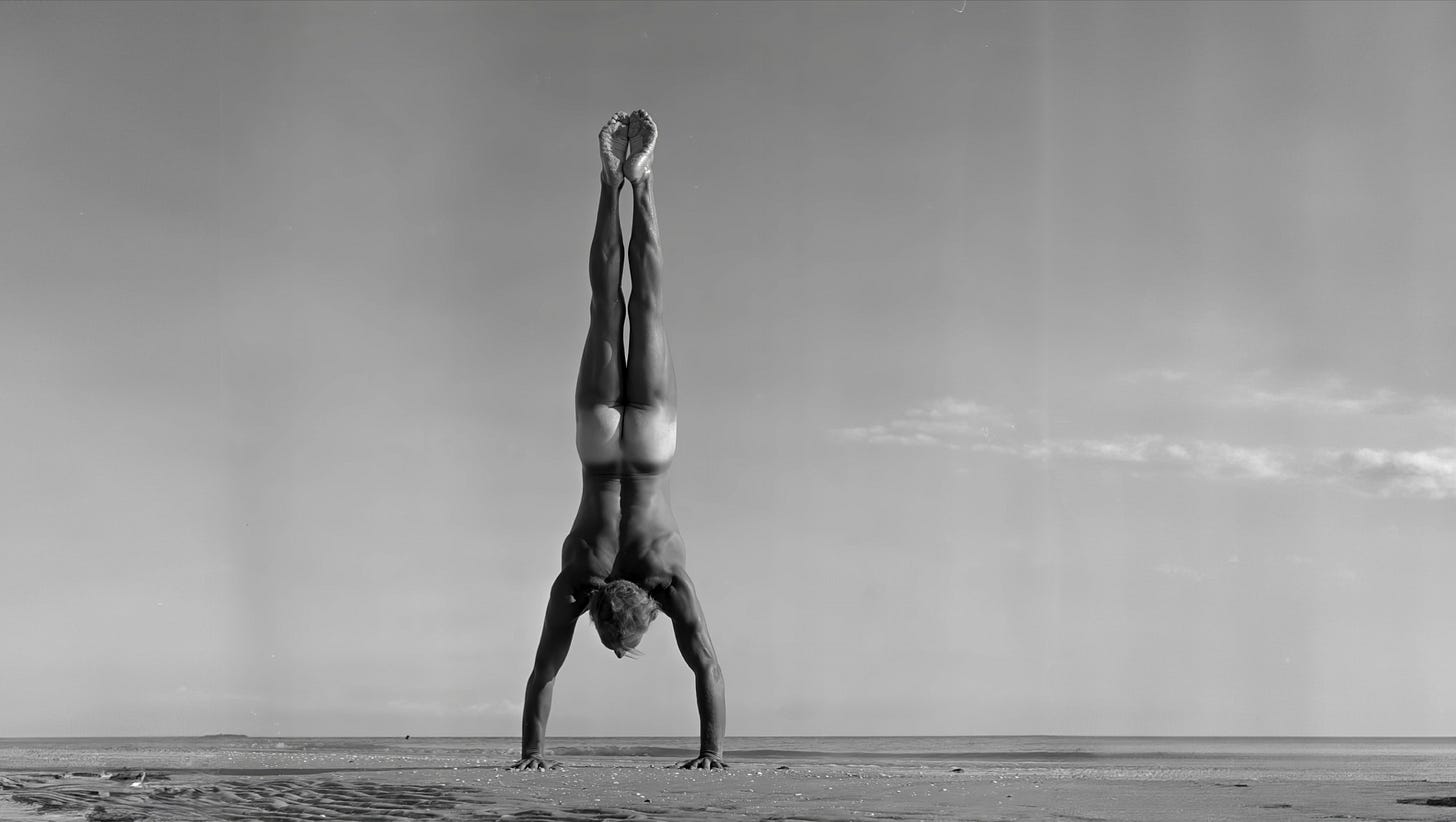 A nude male model performs a perfectly balanced handstand on an empty beach, his body extended straight upward against a wide, cloud-streaked sky, with sand and shoreline stretching out behind him.